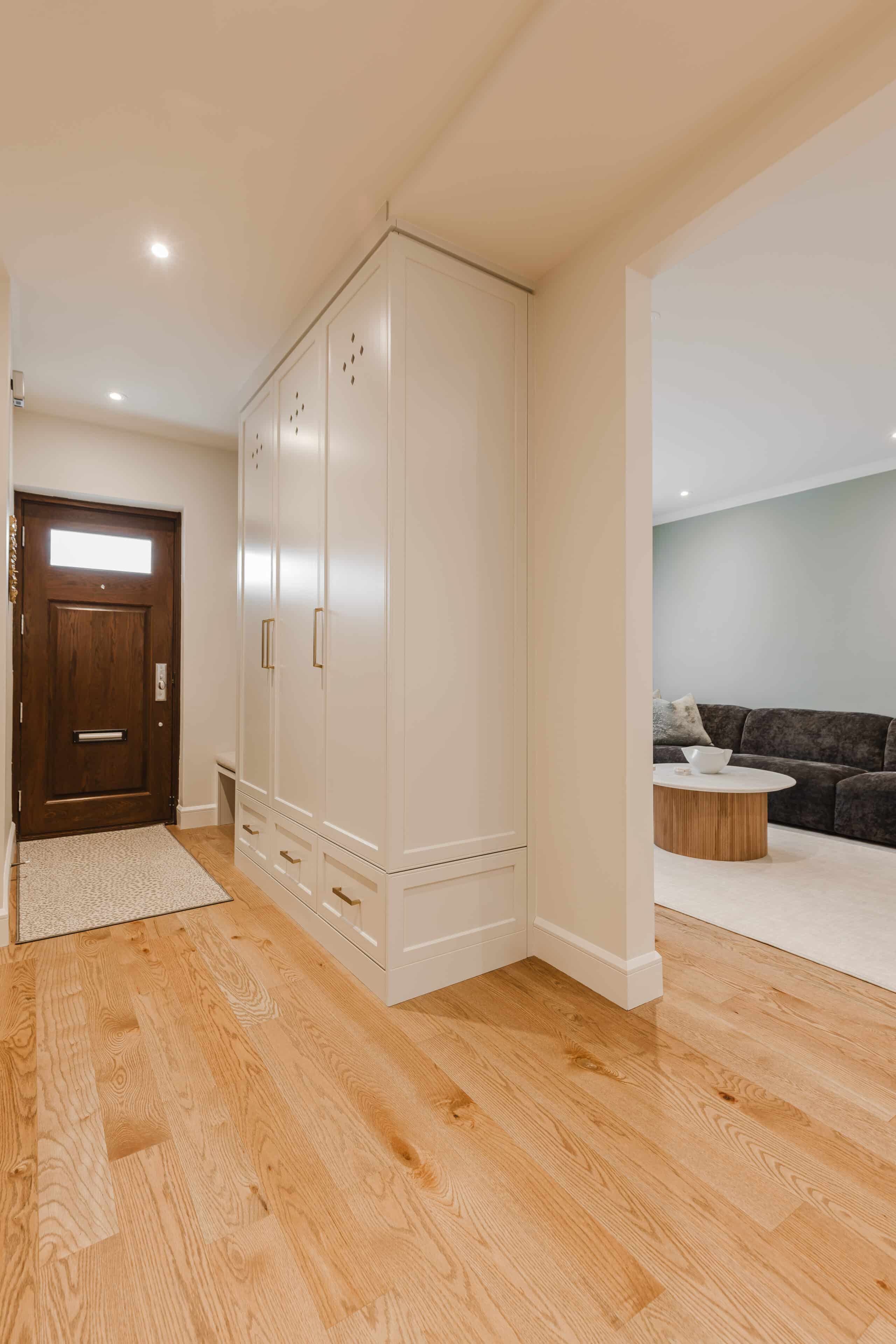 Modern hallway featuring a wooden front door, warm hardwood flooring, and a cozy living area with a dark sectional sofa invites you home.