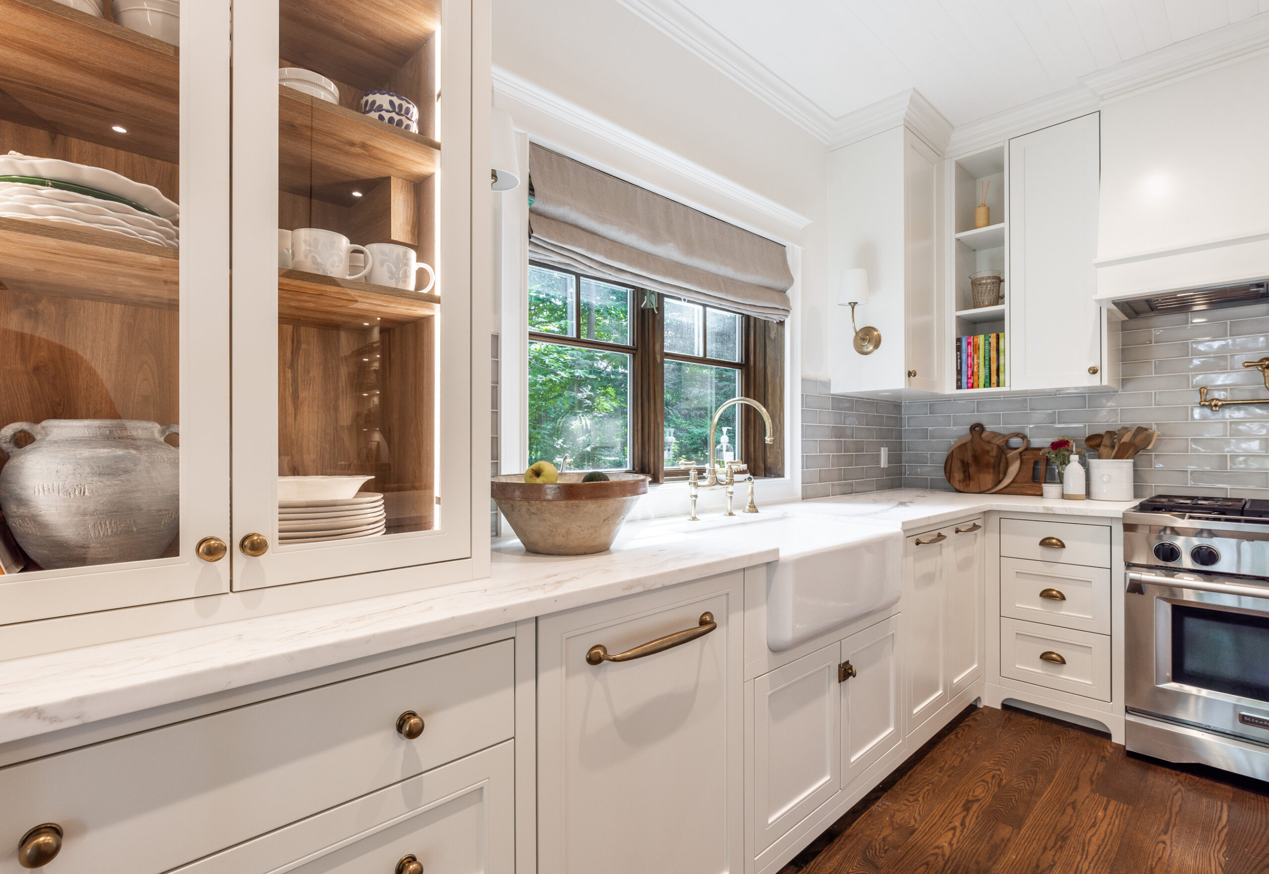 Elegant white kitchen with farmhouse sink, marble countertops, gray subway tiles, and rich wood flooring.