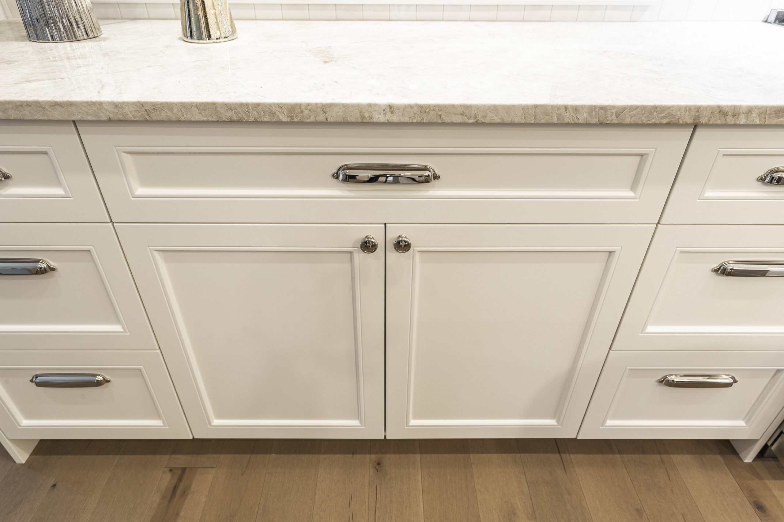 Bright white cabinets and a light stone countertop create a sleek, modern kitchen space with elegant chrome accents and warm hardwood flooring.