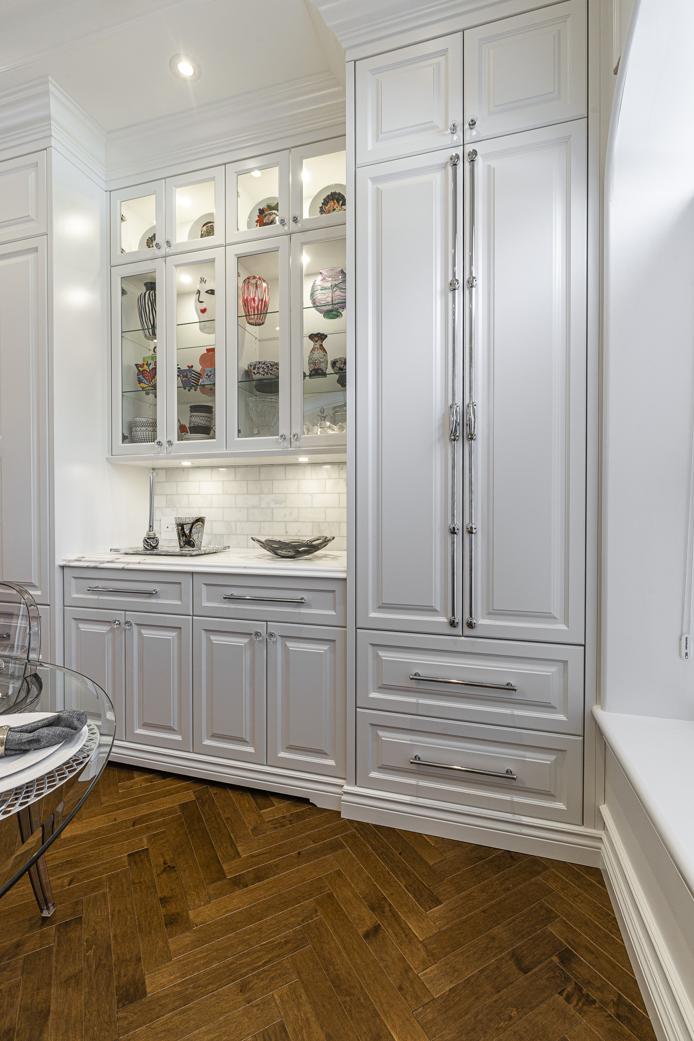 Experience the charm of this elegant white kitchen with glass cabinets, a sleek countertop, and warm herringbone flooring that exudes sophistication.