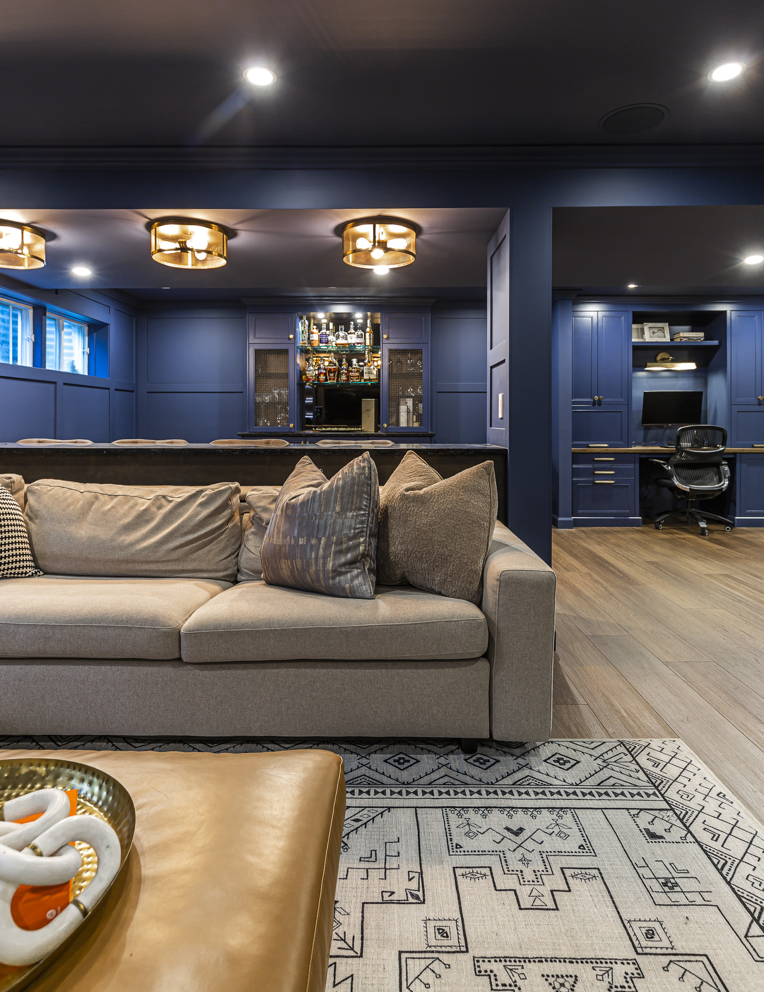 Cozy modern basement retreat with navy walls, plush sofa, chic lighting, and an elegant bar, perfect for relaxation and entertaining.