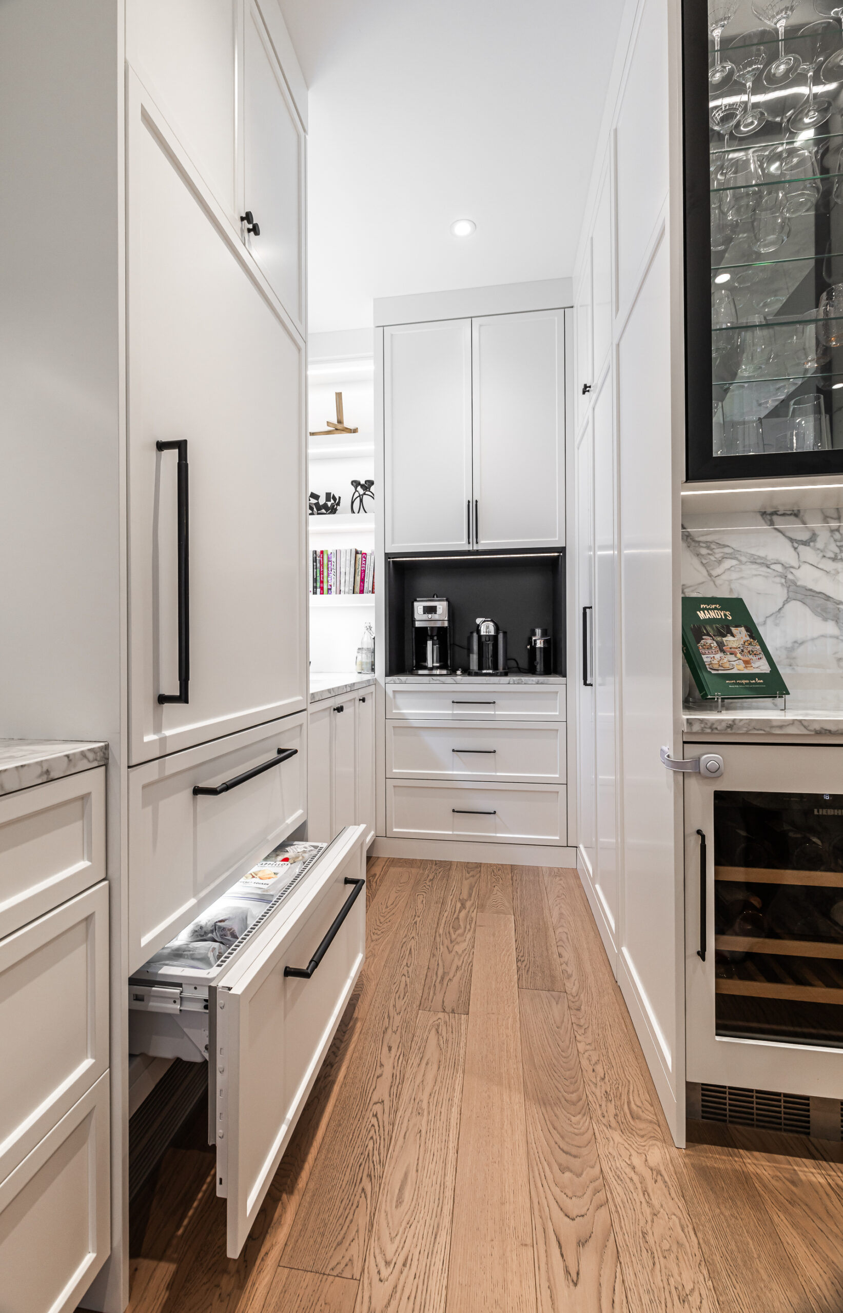 Elegant modern kitchen corridor featuring sleek white cabinets, a coffee station, and a stylish wine fridge complemented by warm wooden flooring.