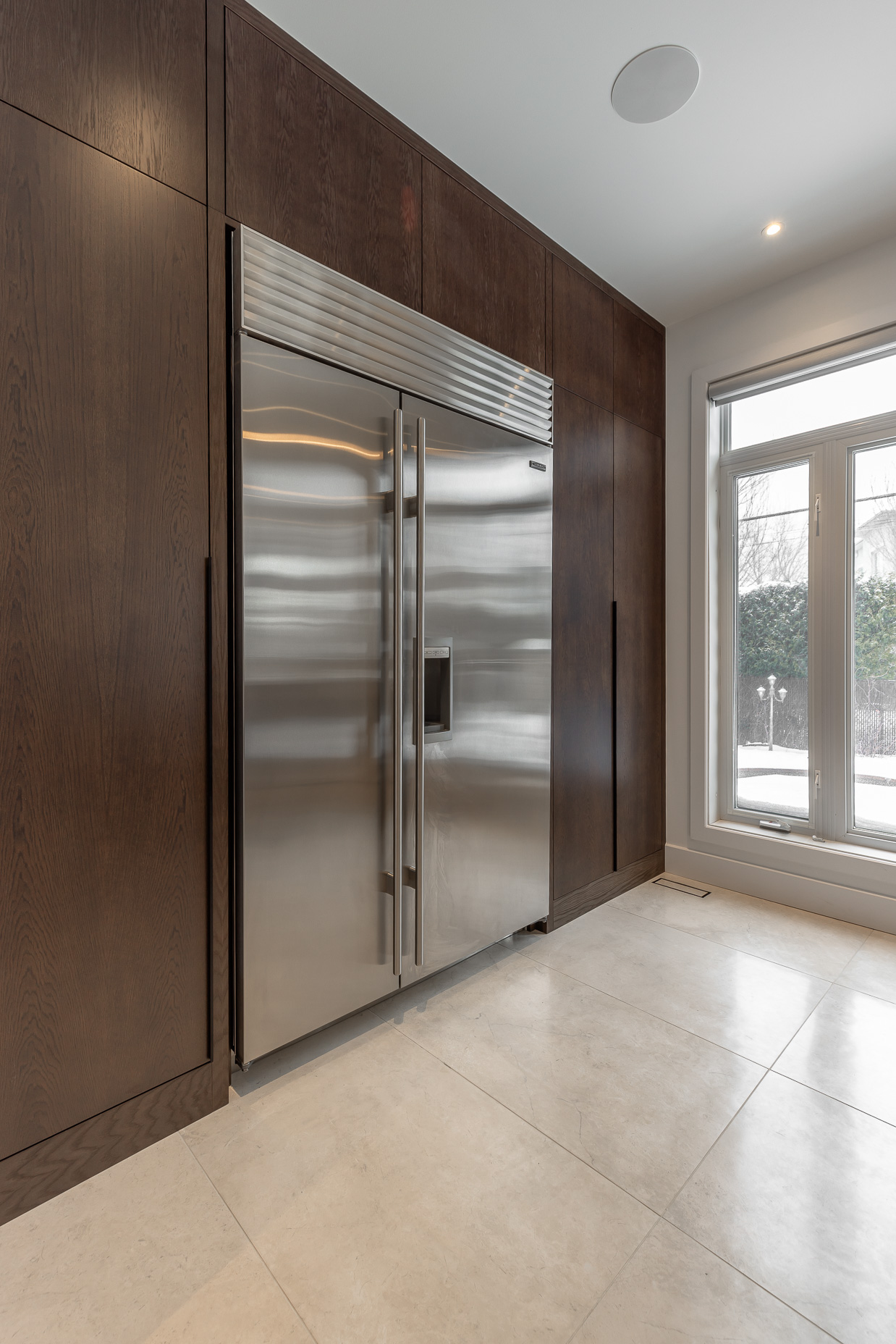 A contemporary kitchen blends a sleek stainless steel fridge with dark wood cabinetry and bright tiles, illuminated by natural light.
