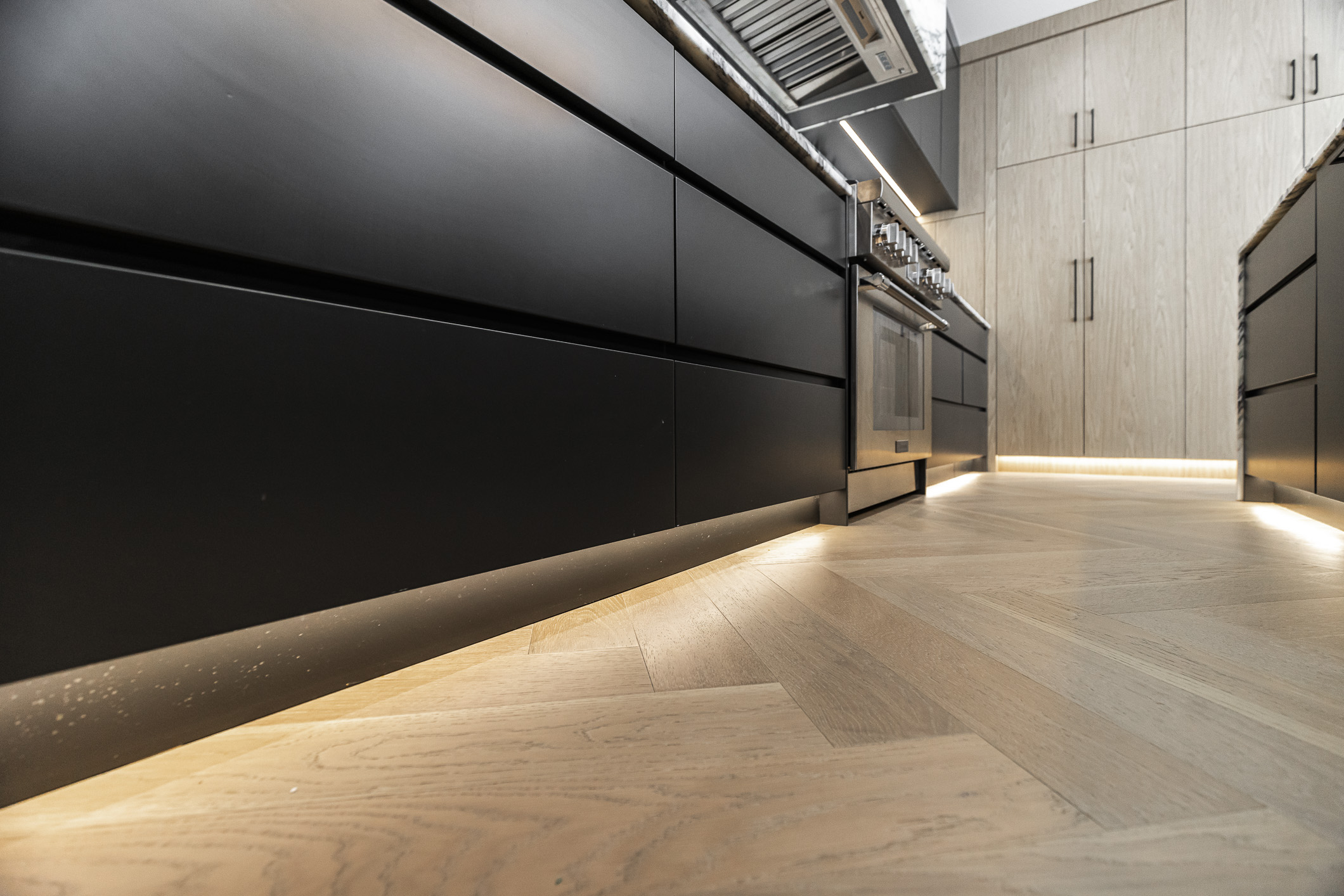 Elegant modern kitchen showcasing contrasting black and light wood cabinetry, ambient lighting, and stylish herringbone flooring for a welcoming ambiance.
