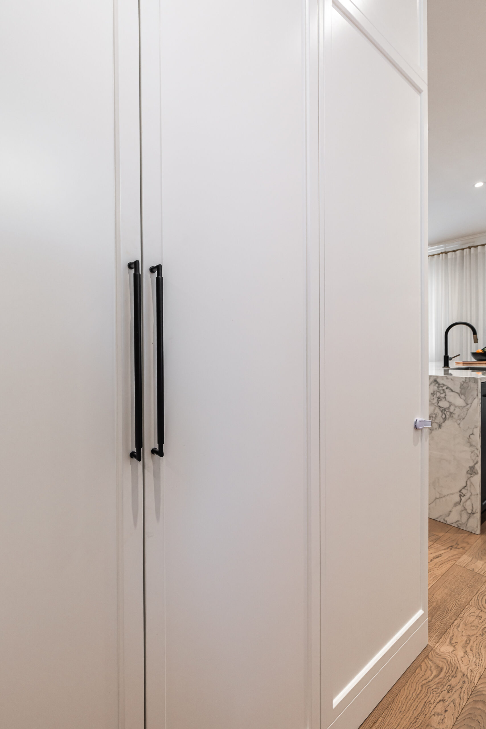 Explore this elegant modern kitchen featuring a seamless white cabinet, striking black hardware, and warm wood flooring for a chic culinary experience.