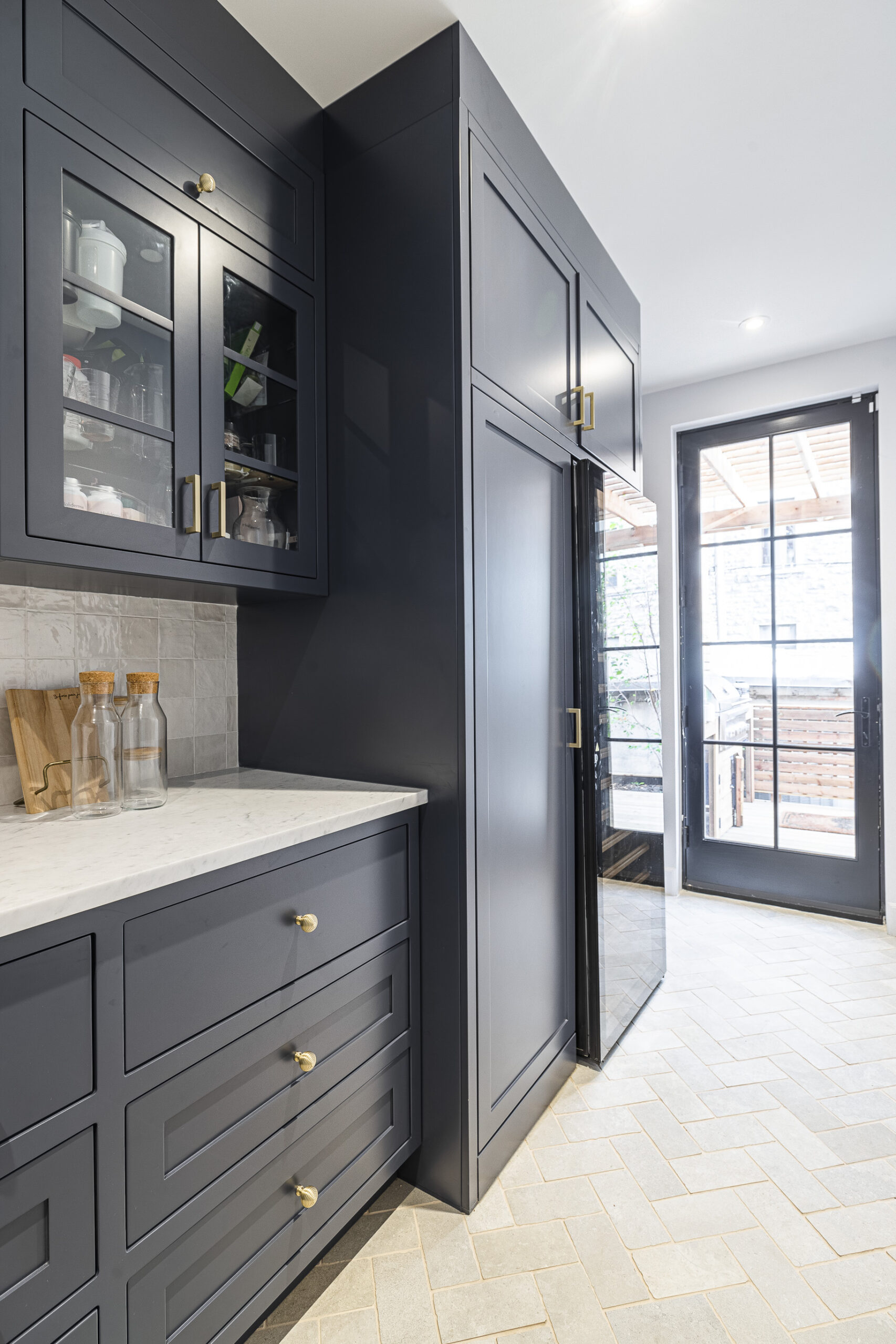 Experience contemporary elegance in this navy blue kitchen featuring glass cabinetry, gold accents, and bright herringbone flooring.