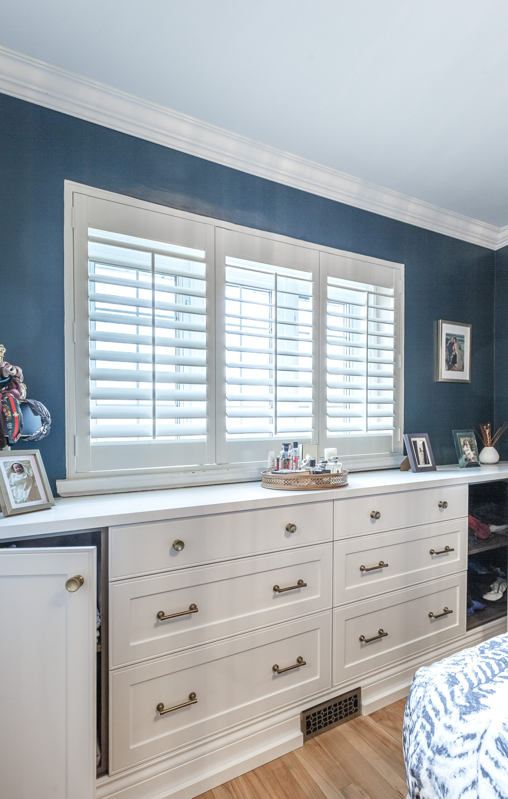 Stylish bedroom featuring creamy cabinetry, deep blue walls, and elegant decor illuminated by natural light through plantation shutters.