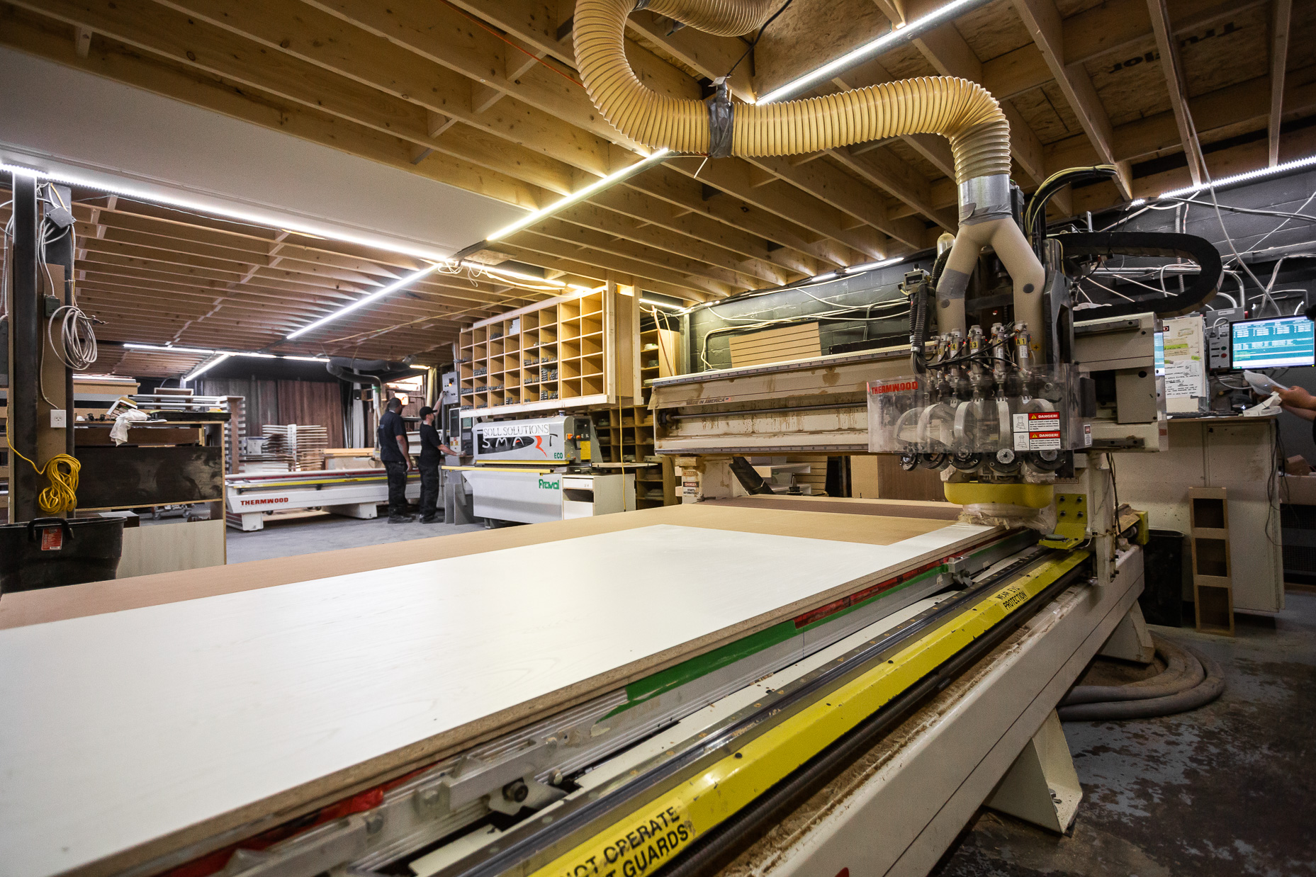 A bustling woodworking shop features a CNC machine precisely cutting material, with craftsmen actively engaged in detailed projects.