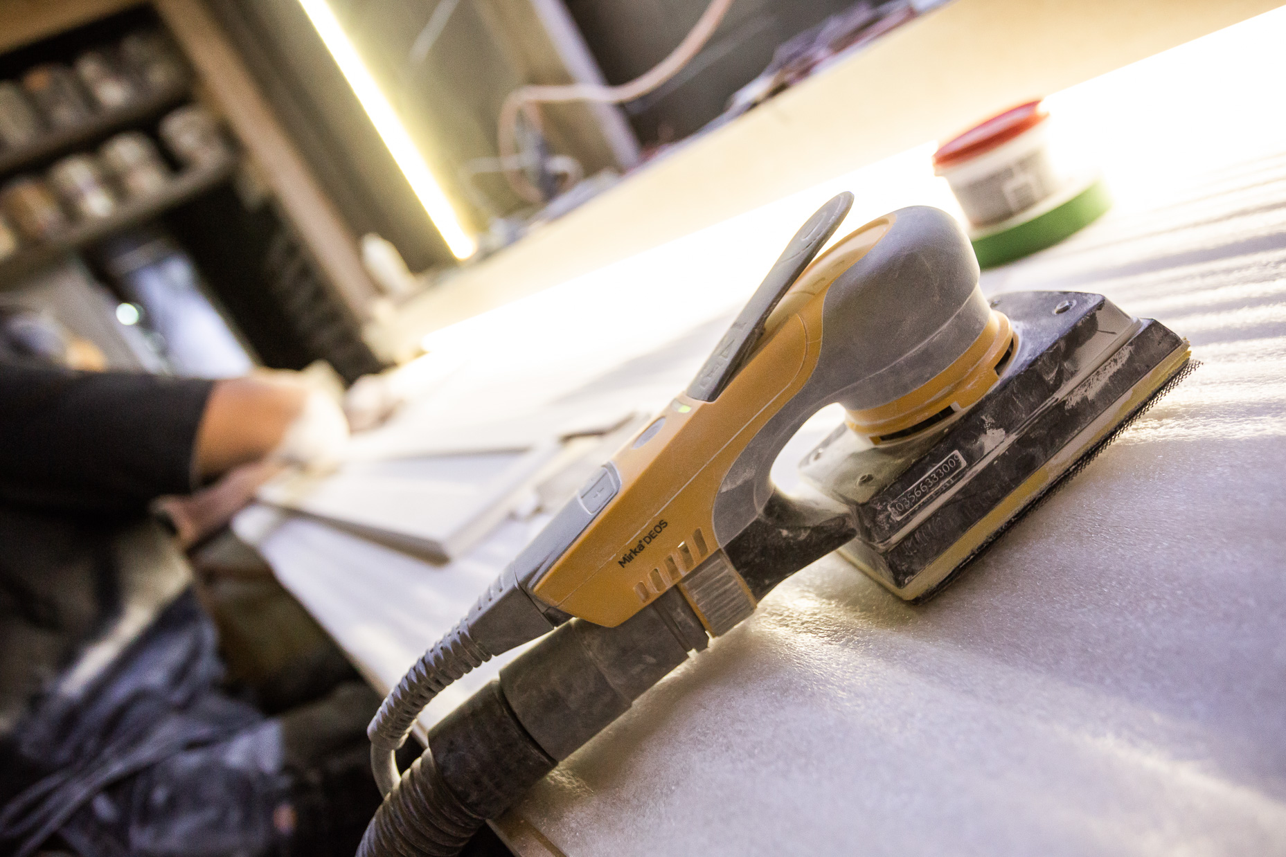 A focused artisan uses a yellow power sander in a well-organized woodworking workspace, surrounded by tools and warm light.