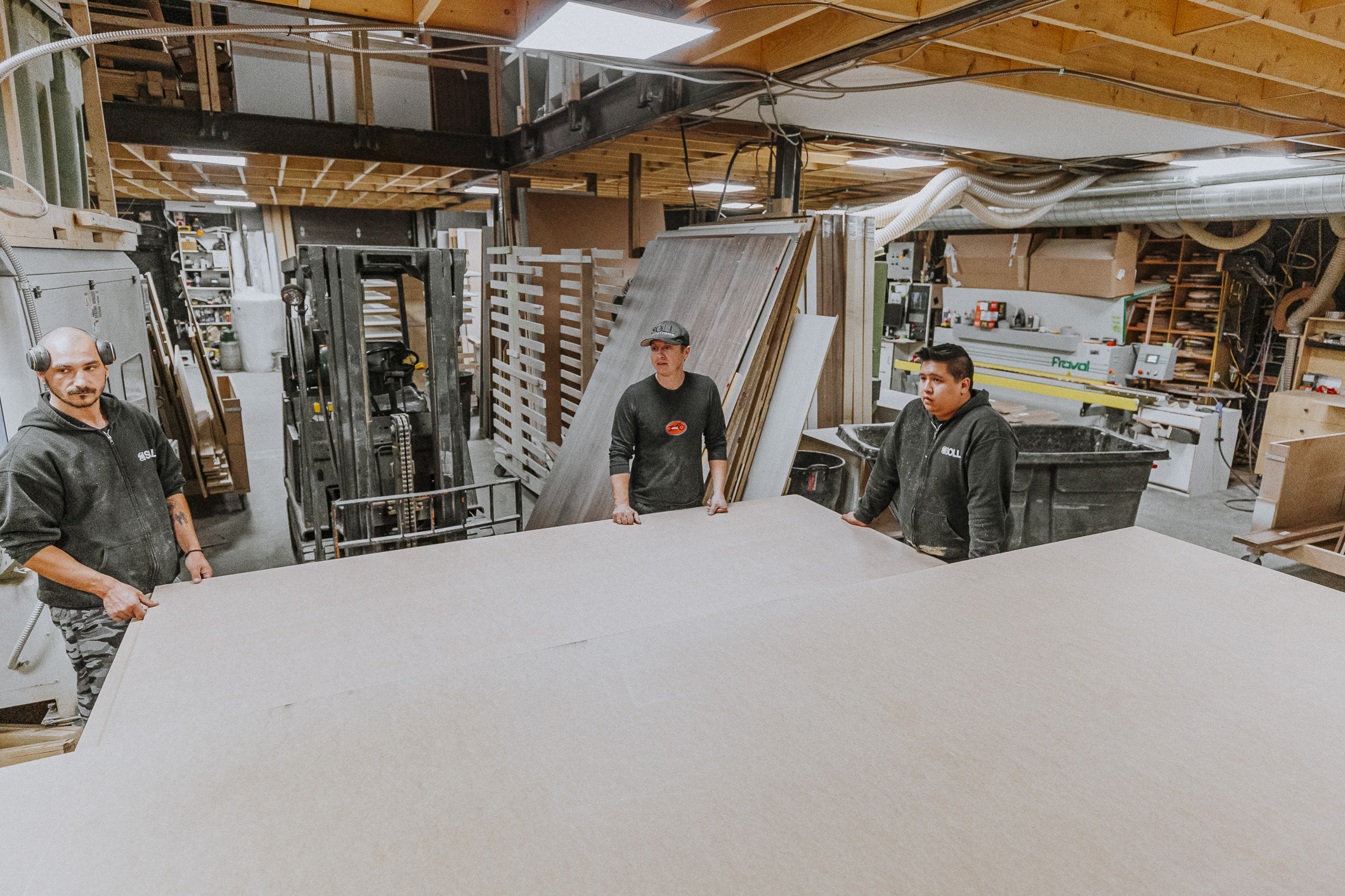 Three workers unite in an industrial workshop to expertly maneuver a large sheet of material, highlighting teamwork and craftsmanship.